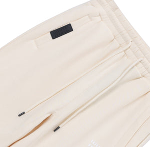 White London Clothing cream cotton jersey joggers with drawstring waist, side pockets, and embossed logo tag on left thigh.